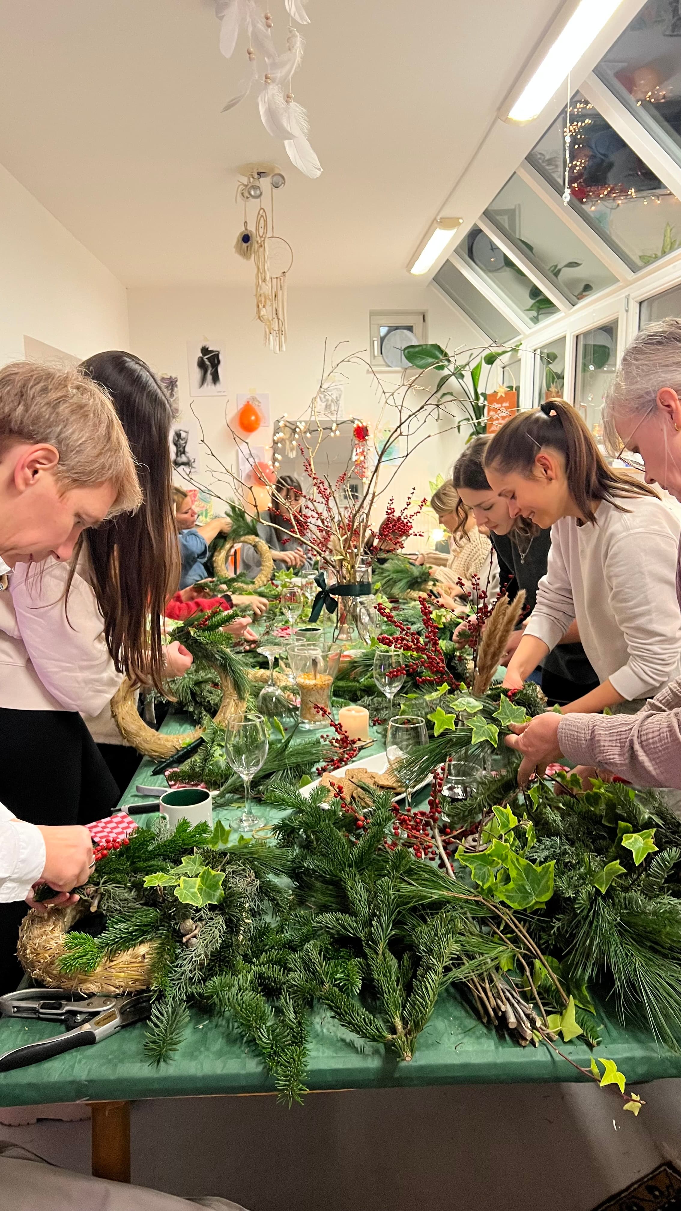 Gruppe arbeitet konzentriert an einem festlich dekorierten Tisch mit Tannenzweigen, Kerzen und Kränzen — gemeinsamer Workshop im Atelier