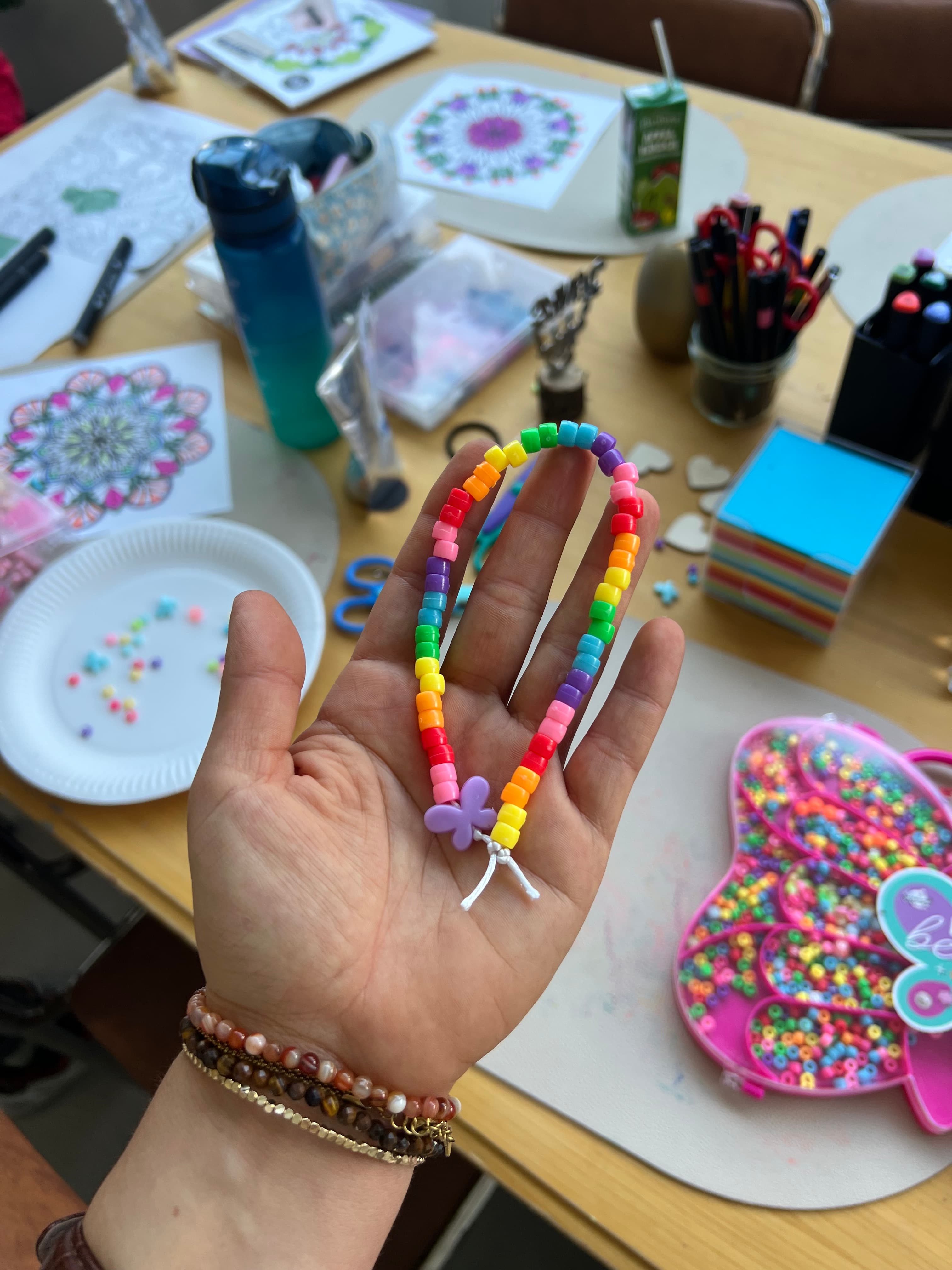 Hand hält ein Regenbogen-Perlenarmband mit lila Schmetterling-Anhänger über einer Mandala-Mal-Station mit Perlentablett