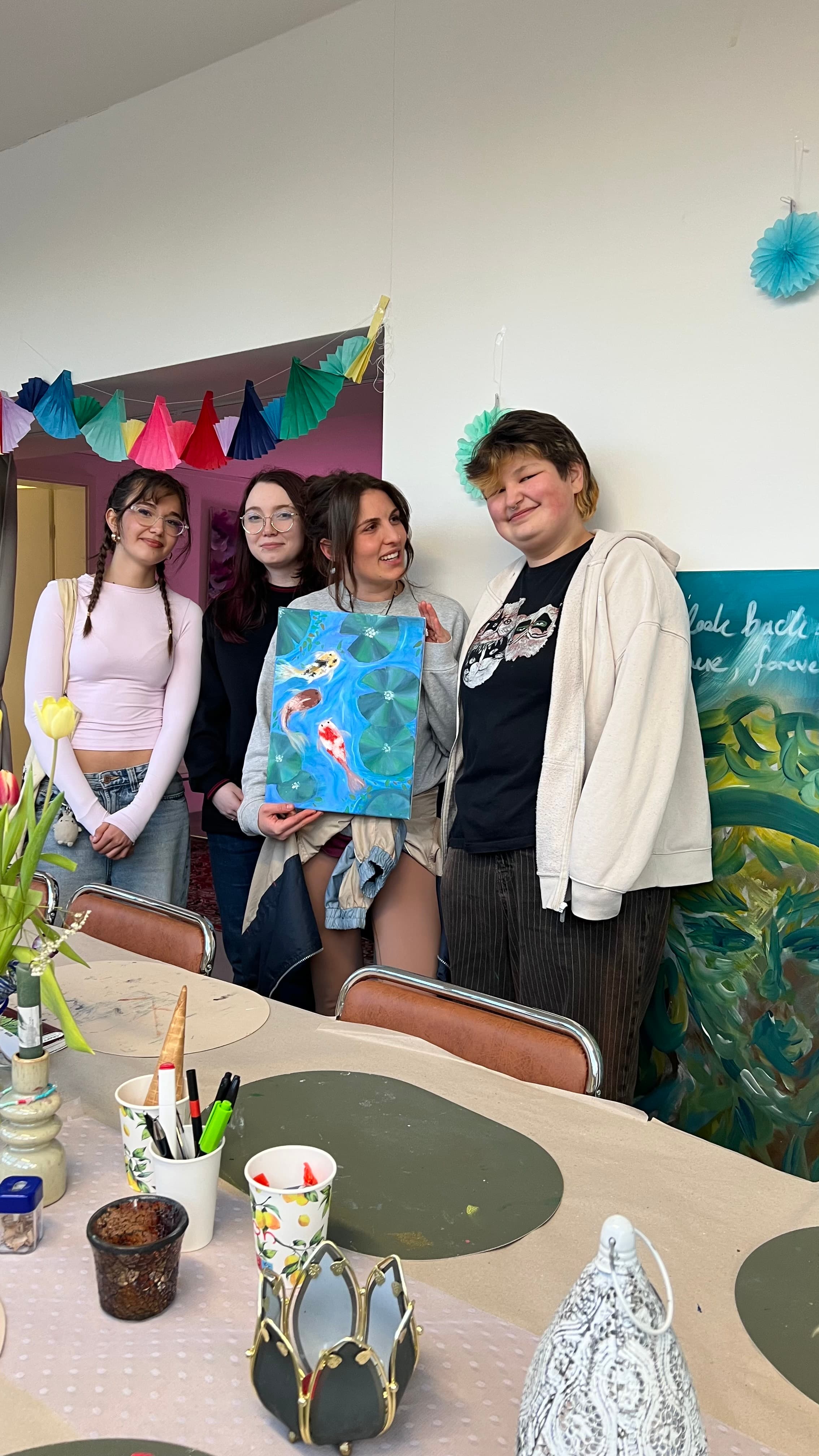 Vier junge Frauen halten gemeinsam ein Aquarell mit Koi-Karpfen-Motiv, dahinter ein festlich dekorierter Tisch mit Tulpen und Papiergirlanden