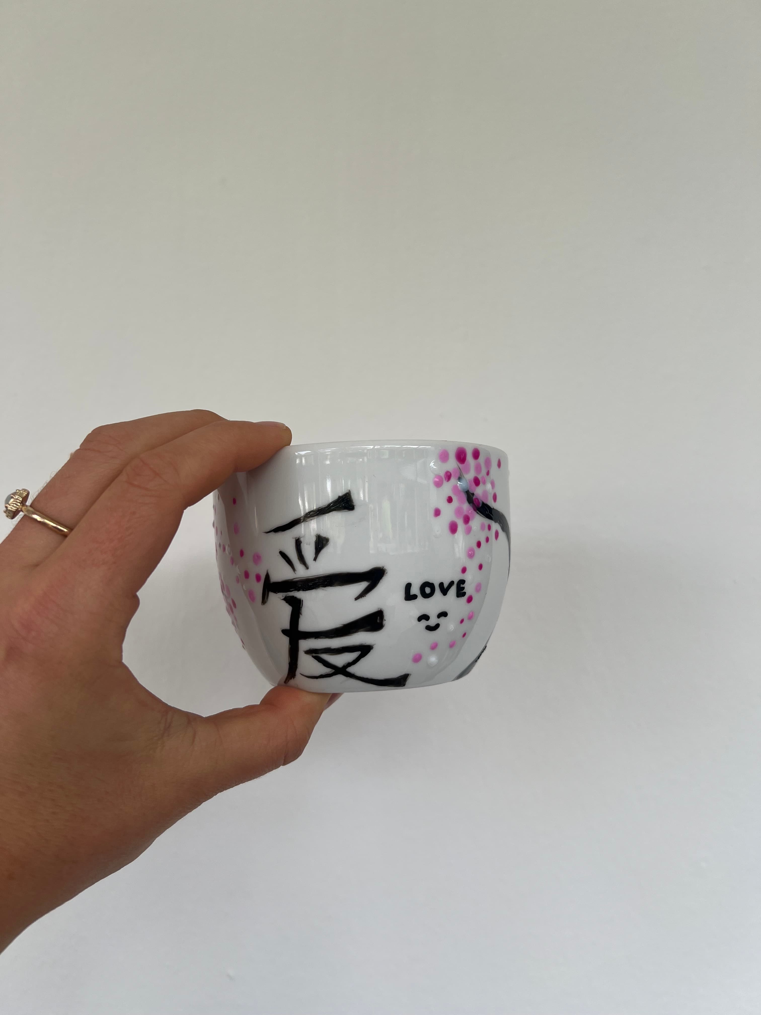 Hand hält eine selbst bemalte Glastasse mit Kirschmotiv und 'LOVE'-Schriftzug in schwarzer Graffiti-Optik vor weißer Wand