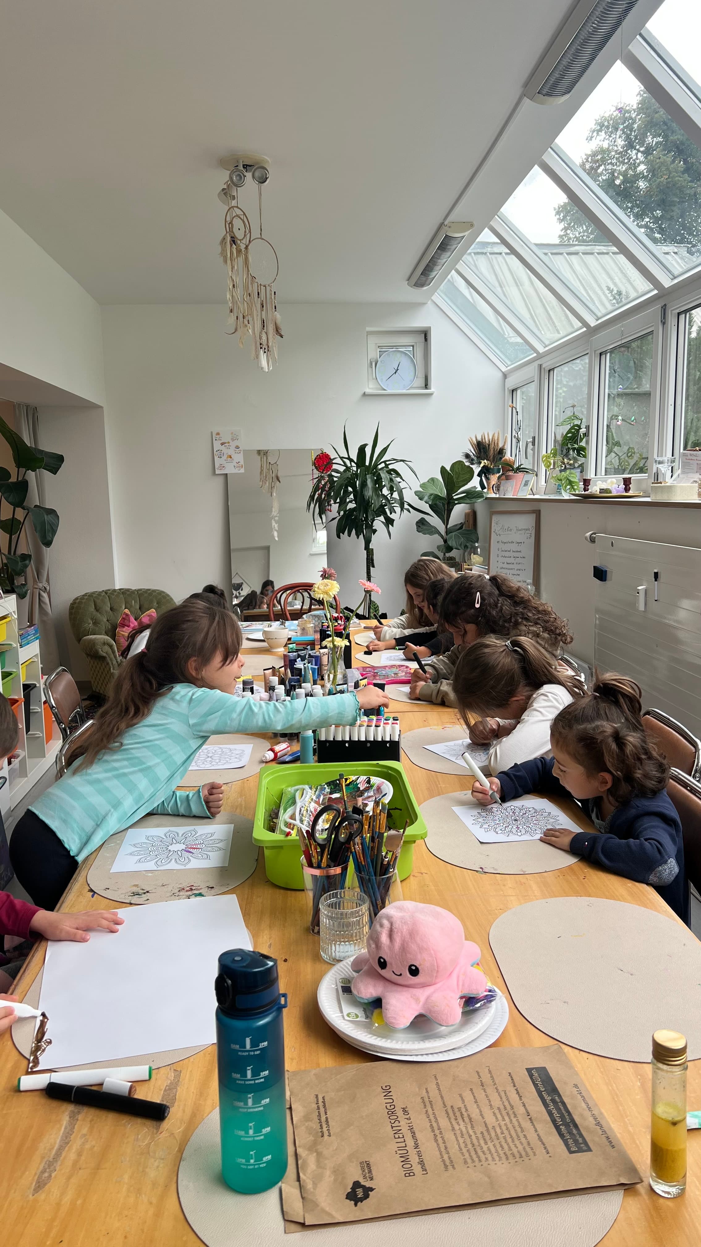 Sonnige Wintergarten-Szene mit mehreren Kindern am langen Tisch beim Mandala-Ausmalen — bunte Stifte und Pflanzen